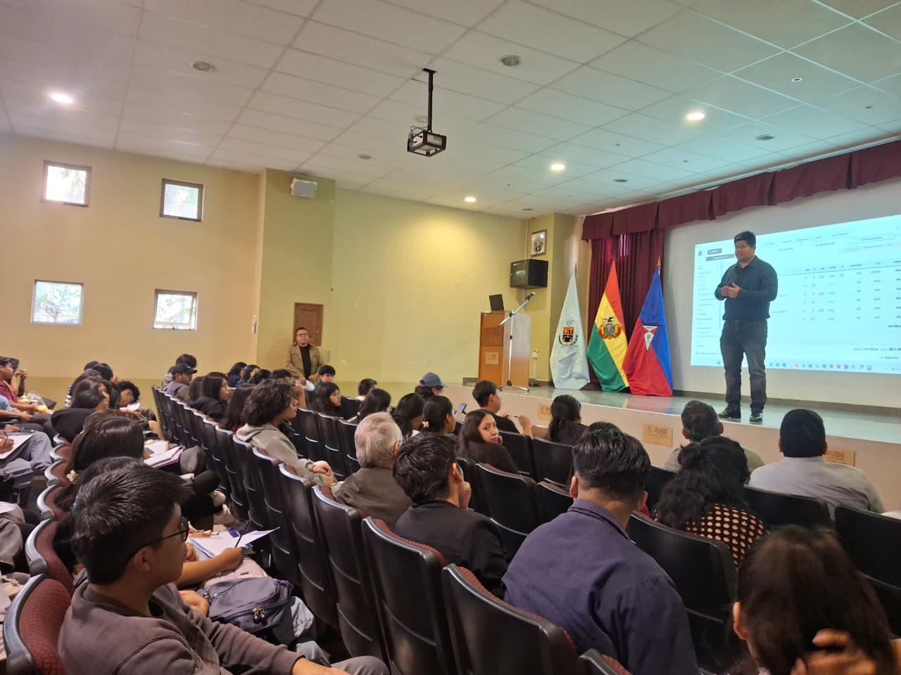 La charla en el auditorio de la Facultad de Arquitectura de la UMSS: el expositor en el escenario, junto a las banderas, con ArqOn proyectado en la pantalla y el público sentado.